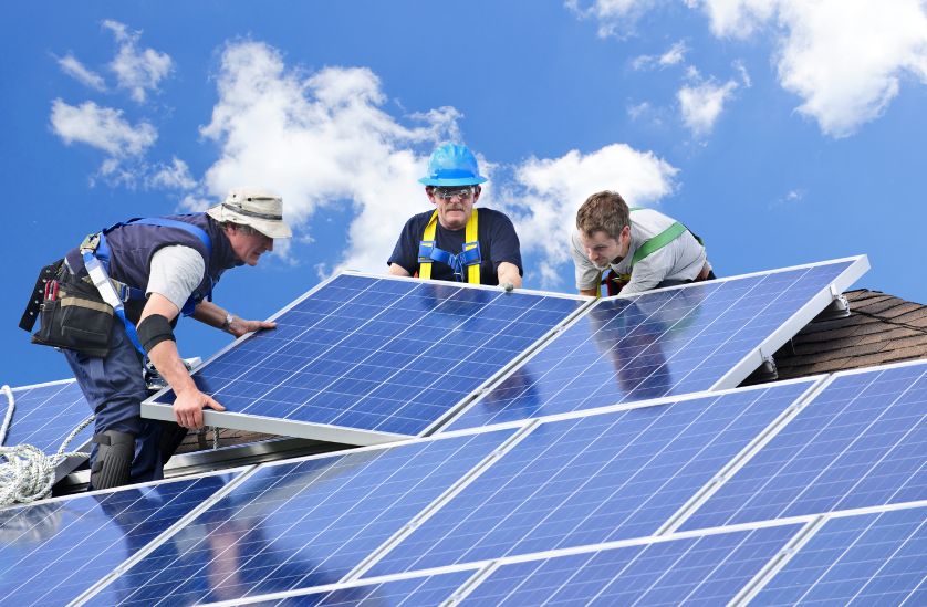 Three workers installing solar panels on a rooftop, showcasing the installation process for renewable energy. The clear blue sky provides a vibrant backdrop, emphasizing the importance of solar energy in sustainable living.